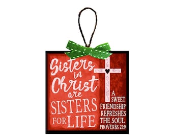 May include: A red wooden ornament with a green ribbon and the text "Sisters in Christ are Sisters for Life" with a cross and the quote "A sweet friendship refreshes the soul Proverbs 27:9"