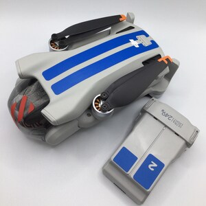 Op de afbeelding: Een witte drone met blauwe race strepen en zwarte propellers. De drone staat op een wit oppervlak naast een grijze batterij met blauwe accenten. De batterij heeft de tekst "Ultra Light 24G" erop gedrukt.