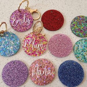 May include: Assortment of round glitter keychains with names written in white. The keychains are in various colours including pink, blue, red, green, and purple.