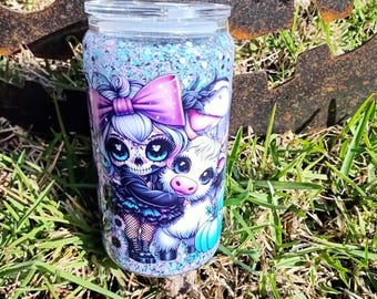 May include: A clear tumbler with a glittery blue and white base. The tumbler features a colourful illustration of a skeleton girl with a pink bow in her hair and a white cow with a pumpkin.