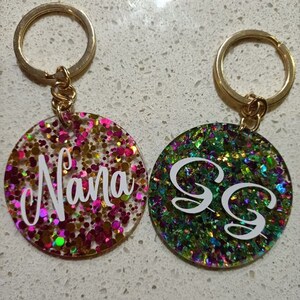 May include: Two gold keychains with round, colourful glitter resin charms. One charm says "Nana" in white lettering on a pink, gold, and green glitter background. The other charm has two white "S" letters on a green, gold, and purple glitter background.