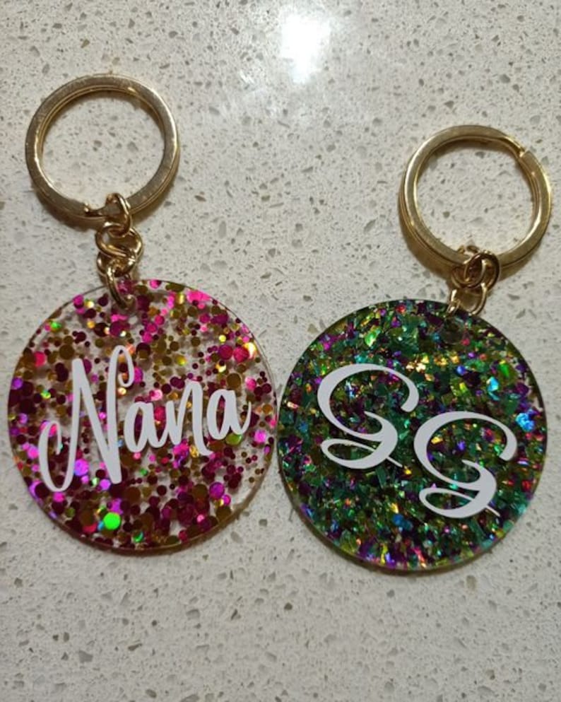 May include: Two gold keychains with round, colourful glitter resin charms. One charm says "Nana" in white lettering on a pink, gold, and green glitter background. The other charm has two white "S" letters on a green, gold, and purple glitter background.
