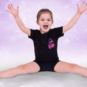 May include: A young girl wearing a black t-shirt with the text "EDN Dance" in pink and blue letters, black shorts, and a big smile, is sitting in a split position with her arms raised in the air. The background is a purple and white sparkly backdrop.
