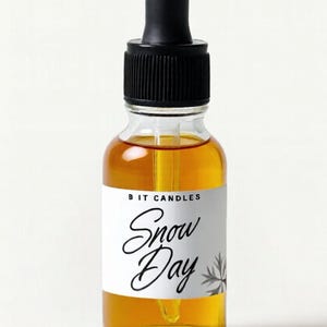 May include: A clear glass bottle with a black dropper and a white label. The label reads "BIT CANDLES Snow Day" in a script font. The bottle contains a golden-colored liquid.