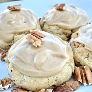 May include: Four pecan cookies with tan frosting are arranged on a white plate. Each cookie is topped with a swirl of frosting and a pecan half. Additional pecan pieces are scattered around the cookies, creating a delicious dessert display.
