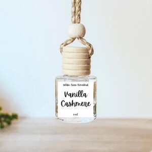 May include: A small clear glass car air freshener bottle with a light wood cap and a thick braided rope hanger. The white label features the text Golden Acres Homestead, Vanilla Cashmere, and 8 ml in black script and sans-serif fonts.