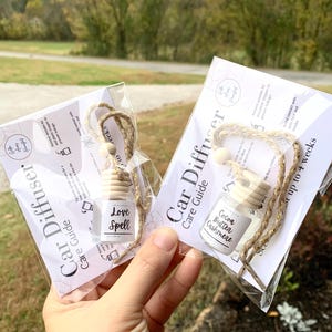 May include: Two car air fresheners in clear glass bottles with wooden tops, hanging from a jute cord. One reads "Love Spell," the other "Cocoa Butter Cashmere." Each is packaged with a care guide.