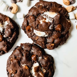 Puede incluir: Tres galletas de chocolate con chispas, malvaviscos, anacardos y chispas de chocolate sobre una superficie blanca.