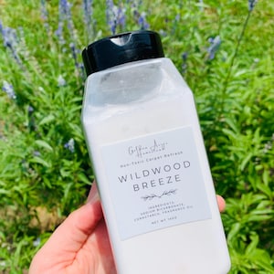 May include: A clear plastic bottle with a black flip-top cap containing white powder. The label reads Wildwood Breeze, Non-Toxic Carpet Refresh, and lists ingredients including sodium bicarbonate, cornstarch, and fragrance oil, with a net weight of 16 oz.