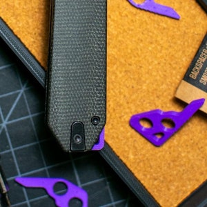 Puede incluir: Un cuchillo de bolsillo negro con mango de fibra de carbono y un botón morado. El cuchillo está en una bandeja negra y gris con una pequeña botella de aceite y un trozo de papel con las palabras "Backspacer M" impresas en él.