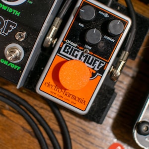 Pode incluir: Um pedal de efeitos Electro-Harmonix Big Muff Pi prateado e preto com um pedal de pé laranja. O pedal tem botões para volume, tom e sustain.