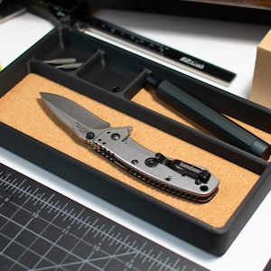 May include: A black organizer tray with a cork liner. The tray contains a silver pocket knife with a black handle and a black pen.
