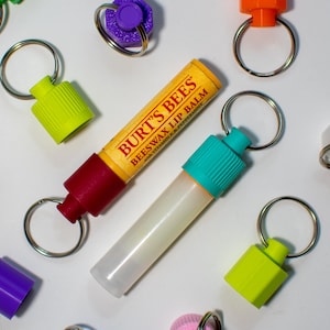May include: A Burt's Bees Beeswax Lip Balm tube attached to a silver key ring with a green cap. The lip balm is next to a clear bottle with a turquoise cap, also attached to a silver key ring. There are several other key rings with colorful caps in the background.