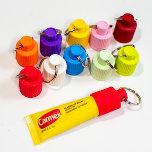Könnte beinhalten: Eine gelbe Tube Carmex Classic Lip Balm Medicated mit rotem Deckel und silbernem Schlüsselring. Die Tube ist umgeben von zehn bunten Schlüsselanhänger-Flaschenverschlüssen in verschiedenen Farben, darunter Rot, Orange, Gelb, Grün, Blau, Lila, Rosa und Weiß.
