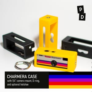 以下が含まれることがあります： 銀色のキーホルダーリングが付いた黄色い保護ケースに入ったKodak Charmeraカメラ。ケースには1/4インチのカメラマウントとオプションのホットシューが付いています。背景には黒と黄色の空のケースが見えます。