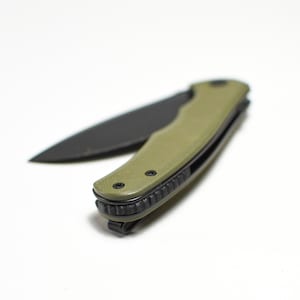 May include: A black folding knife with a green handle. The handle has a textured surface and a black clip.