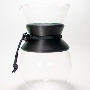 May include: A clear glass coffee carafe with a black rubber band and a black plastic stopper. The carafe has a wide mouth and a narrow spout.