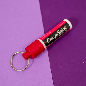 Può includere: Un balsamo per labbra ChapStick rosso attaccato a un portachiavi argentato. Il tubo del balsamo per labbra ha una scritta bianca che recita "ChapStick Skin Protectant".