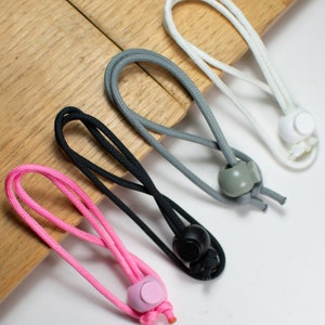May include: Four different colored cords with adjustable toggles. The cords are pink, black, gray, and white. The toggles are made of plastic and are in the colors black, gray, white, and pink.