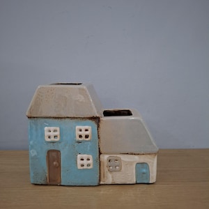 Può includere: Un vaso da fiori in ceramica a forma di due piccole case, una blu e una bianca, con porte e finestre marroni. Le case sono unite e hanno un aspetto rustico e fatto a mano.