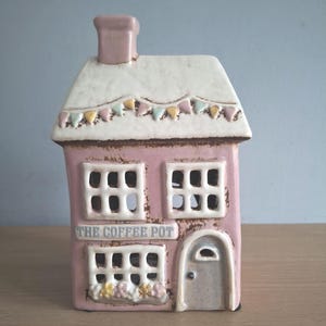 May include: A pink ceramic house-shaped candle holder with a white roof and chimney. The house features windows, a door, and the text "THE COFFEE POT". The roof is decorated with small, colourful flags.