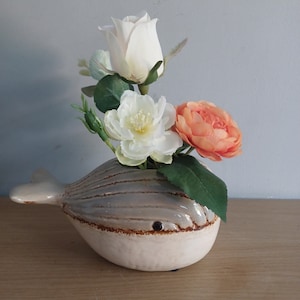 May include: A ceramic whale-shaped vase in cream and gray, holding a bouquet of artificial flowers. The flowers include a white rose, a white peony, and an orange peony, with green leaves. The vase is on a wooden surface.