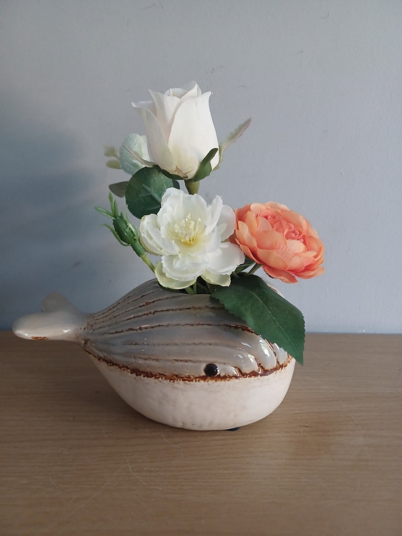 May include: A ceramic whale-shaped vase in cream and gray, holding a bouquet of artificial flowers. The flowers include a white rose, a white peony, and an orange peony, with green leaves. The vase is on a wooden surface.