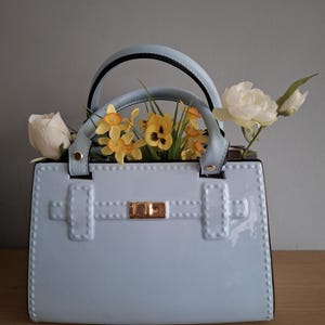 Può includere: Una borsa a mano azzurra lucida, con chiusura e manico dorati. La borsa è piena di fiori gialli e bianchi, tra cui rose e narcisi, creando un'estetica primaverile.