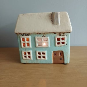 May include: A ceramic house-shaped candle holder with a light blue body and a white roof. The house has six square windows that glow with light, and a brown door. The candle holder is a decorative item.