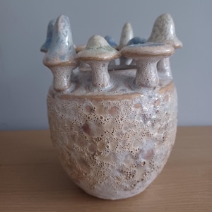May include: A ceramic vase with a mushroom design. The vase is white with brown and blue accents. The mushrooms are arranged in a circle around the top of the vase.