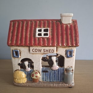 May include: A ceramic cow shed figurine featuring a red roof, white walls, and blue doors. Two black and white cows peek out from the stalls. The structure includes a chimney, a small window, a yellow hay bale, a potted plant, and a grey milk churn.