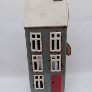 May include: A ceramic house shaped candle holder with a white roof, blue walls, and red door. The house has three windows on each side and a small chimney on top.