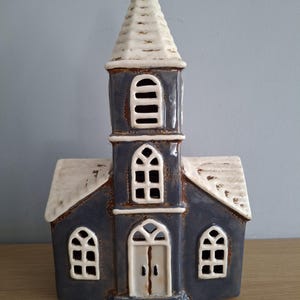 May include: A ceramic church ornament with a steeple and arched windows. The church is painted in a dark blue with white accents on the roof, steeple, and window frames. The doors are white. The ornament is on a wooden surface.