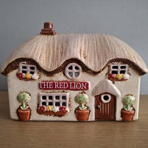 May include: A ceramic cottage figurine with a thatched roof, windows, and a door. The sign reads "THE RED LION". The cottage has potted plants and decorative details. The color palette includes beige, brown, red, and green.