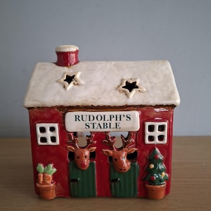 Könnte beinhalten: Eine hausförmige Keramikfigur mit roter Basis und weißem Dach, die den Schriftzug "RUDOLPH'S STABLE" über zwei grünen Stalltüren trägt. Zwei Rentierköpfe schauen aus den Türen hervor, flankiert von einer Topfpflanze mit Karotten und einem Tannenbaum.