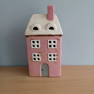 May include: A pink ceramic house with white trim and a gray door. The house has two windows on each floor and two small round windows on the roof.