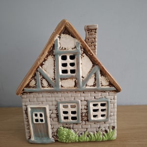 Può includere: Statuina a forma di casa in ceramica con tetto marrone, pareti a forma di mattoni e piccole finestre. La casa ha un camino e un piccolo cespuglio verde alla base. Le finiture sono azzurre e bianche.