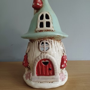 Può includere: Una stravagante casetta delle fate in ceramica con tetto azzurro e dettagli a forma di fungo rosso. La casa ha una porta rossa con un ritaglio a forma di cuore e piccole finestre. La casa è dipinta di bianco e crema.