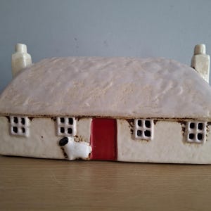 May include: A white ceramic cottage with a red door and a small white sheep figurine in front. The roof is textured to look like thatch.