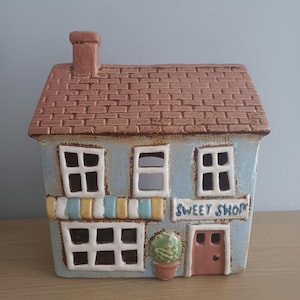 May include: A ceramic model of a sweet shop with a light blue exterior, a brown tiled roof, and a chimney. The shop features multiple windows, a door, and a sign that reads "SWEET SHOP". A small potted plant is beside the door.
