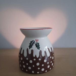 Op de afbeelding: Een keramische oliebrander in de vorm van een kerstpudding. De bovenkant is wit met een rode rand en de onderkant is bruin met witte stippen. Groene hulst en rode bessen versieren de voorkant. Het staat op een houten oppervlak.