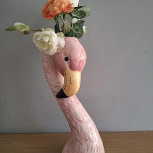 May include: A ceramic flamingo-shaped vase with a glossy pink finish and textured feather details. The flamingo has a curved neck, a yellow beak with a black tip, and a black eye. Artificial white and orange flowers are arranged inside the top.