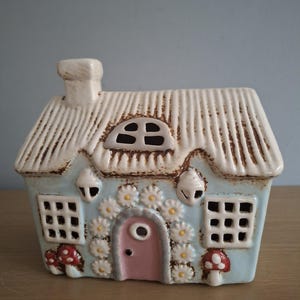 Può includere: Un cottage in ceramica azzurra con tetto di paglia bianco e porta rosa. Il cottage è decorato con fiori a forma di margherita e accenti di funghi rossi. Finestre e un camino completano il design stravagante.