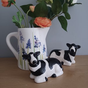 May include: Two black and white ceramic cow salt and pepper shakers with a white jug with blue flowers on it. The jug has a bouquet of orange and white flowers in it.