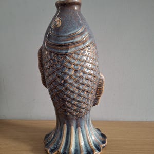 Op de afbeelding: Een keramische vaas in de vorm van een vis, met een blauw en bruin glazuur. De vaas heeft gedetailleerde schubben, vinnen en een uitlopende basis. Een decoratief item.