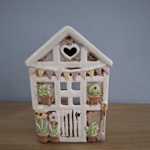 May include: A ceramic house ornament with a heart-shaped window and a small bunting. The house is decorated with potted plants, flowers, and a miniature shovel. The colors are white, brown, and pastel shades.