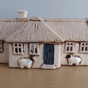 May include: Ceramic cottage ornament with a thatched roof, chimney, and small windows. The off-white house features a dark blue door and two sheep figurines on the front. The cottage is a decorative item.