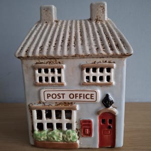 Può includere: Statuetta in ceramica dell'ufficio postale a forma di casa. L'edificio grigio chiaro ha un tetto strutturato, due camini e una porta rossa. L'insegna recita "POST OFFICE". Sono visibili anche finestre e una cassetta postale.