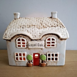 Op de afbeelding: Een keramische kandelaar in de vorm van een huis met een wit dak en lichtblauwe muren. De ramen en deuren zijn verlicht en het bordje luidt "Royal Oak". Twee kleine potbomen flankeren de rode deur.