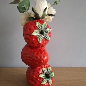 May include: A whimsical, stacked strawberry vase in bright red ceramic, topped with white roses and greenery. The vase is designed to resemble three strawberries, each with a green and cream-colored flower detail. A charming decorative piece.